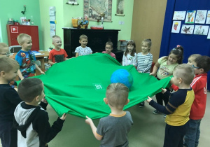 Dzieci z grupy Motylki trzymają duży kawałek materiału na którym znajduje się duża piłka. Unoszą i opuszczają materiał w taki sposób żeby piłka krążyła „Dookoła świata”