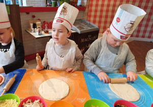 Trzech chłopców wałkuje placek na pizzę, natomiast jeden chłopiec posypuje placek żółtym serem.