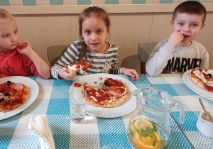 Dwie dziewczynki i chłopiec jedzą własnoręcznie przygotowaną pizzę.