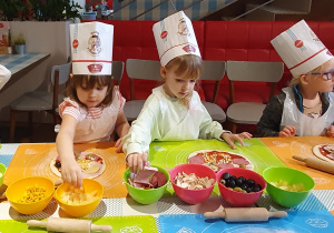 Troje dzieci prezentuje przygotowane pizze, natomiast dwie dziewczynki układają na pizzy ananasa i szynkę.