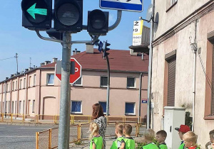 Dzieci z nauczycielką czekają na przejściu dla pieszych na zielone światło sygnalizatora.
