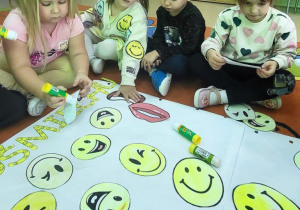 Dziewczynka przykleja wyciętą z papieru uśmiechniętą buzię na karton.