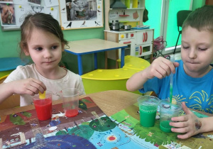 Dziewczynka z chłopcem przelewają wodę z kubeczka do kubeczka z użyciem pipet.